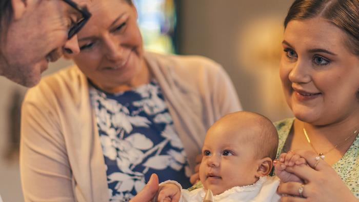 Ett dop i en kyrka. En kvinna med sin man bakom sig, håller barnet i famnen. En manlig präst håller barnets hand.