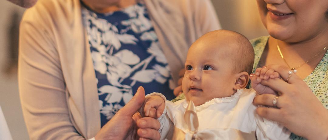 Ett dop i en kyrka. En kvinna med sin man bakom sig, håller barnet i famnen. En manlig präst håller barnets hand.