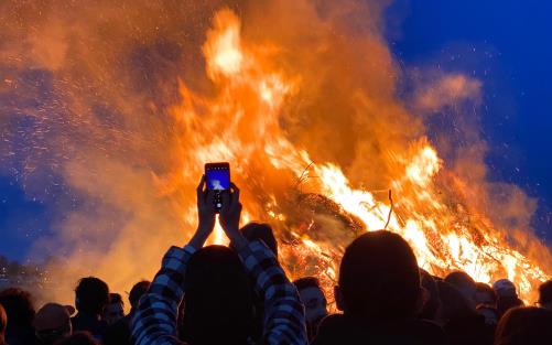 En grupp människor vid en stor majbrasa. Någon håller upp sin mobiltelefon och tar bild.