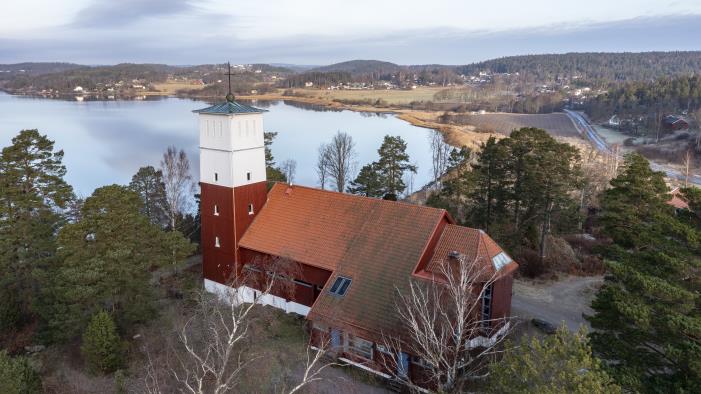 Drönarbild på en faluröd kyrka med vitt torn tegeltak, vackert belägen vid en sjö.