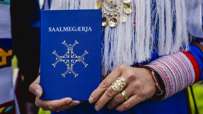 En person i samiska kläder håller fram en samisk psalmbok.
