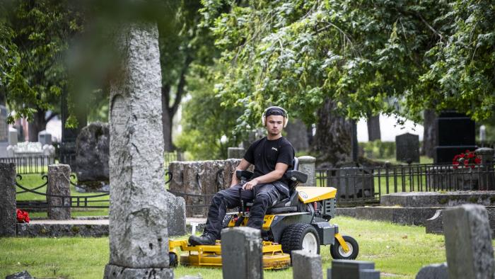 En ung kille klipper gräset mellan gravarna med en åkgräsklippare på en kyrkogård.