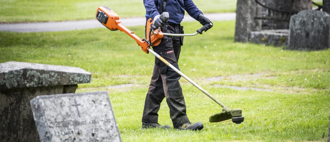 En person går med en grästrimmer mellan gravarna på en kyrkogård.