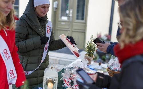 Två kvinnor med Act Svenska kyrkan banderoller över axeln säljer lotter och kakor på julmarknad.