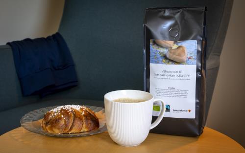 En kaffekopp, en kanelbulle och en påse med kaffe från Svenska kyrkan står på ett bord.