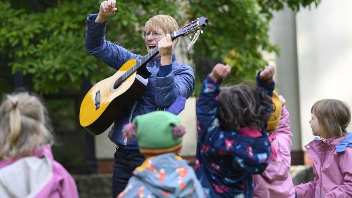 En kvinna står utomhus och spelar gitarr för en grupp barn.