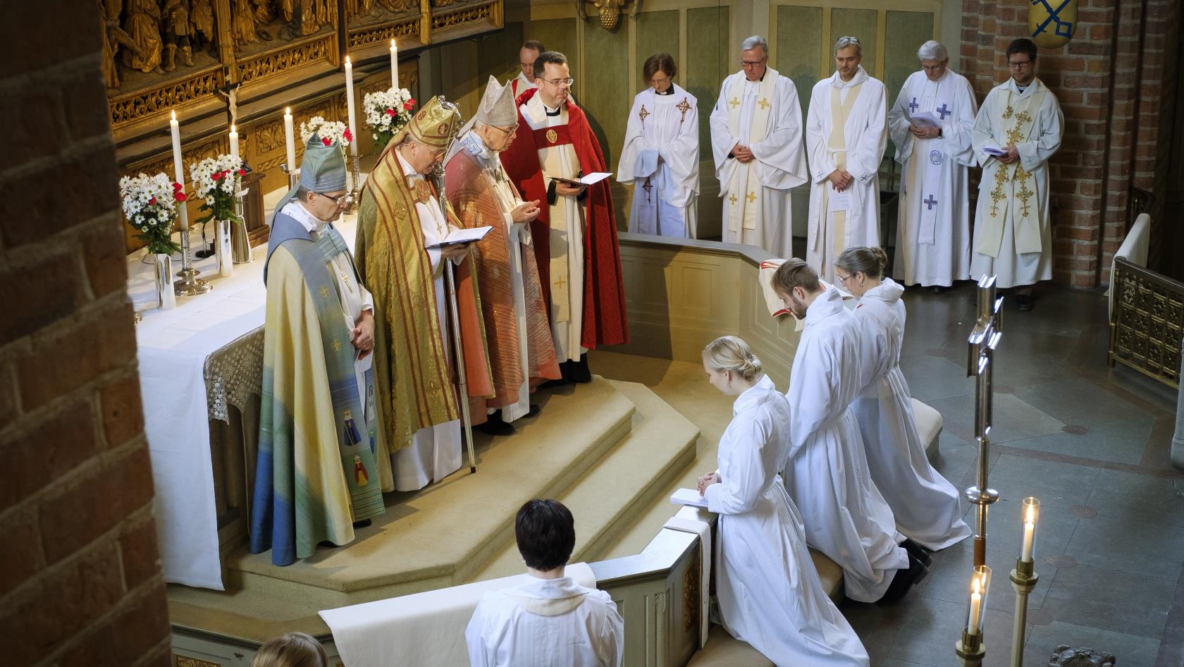 Fyra biskopar står vid ett altare, vända mot tre vitskrudade personer som knäböjer.