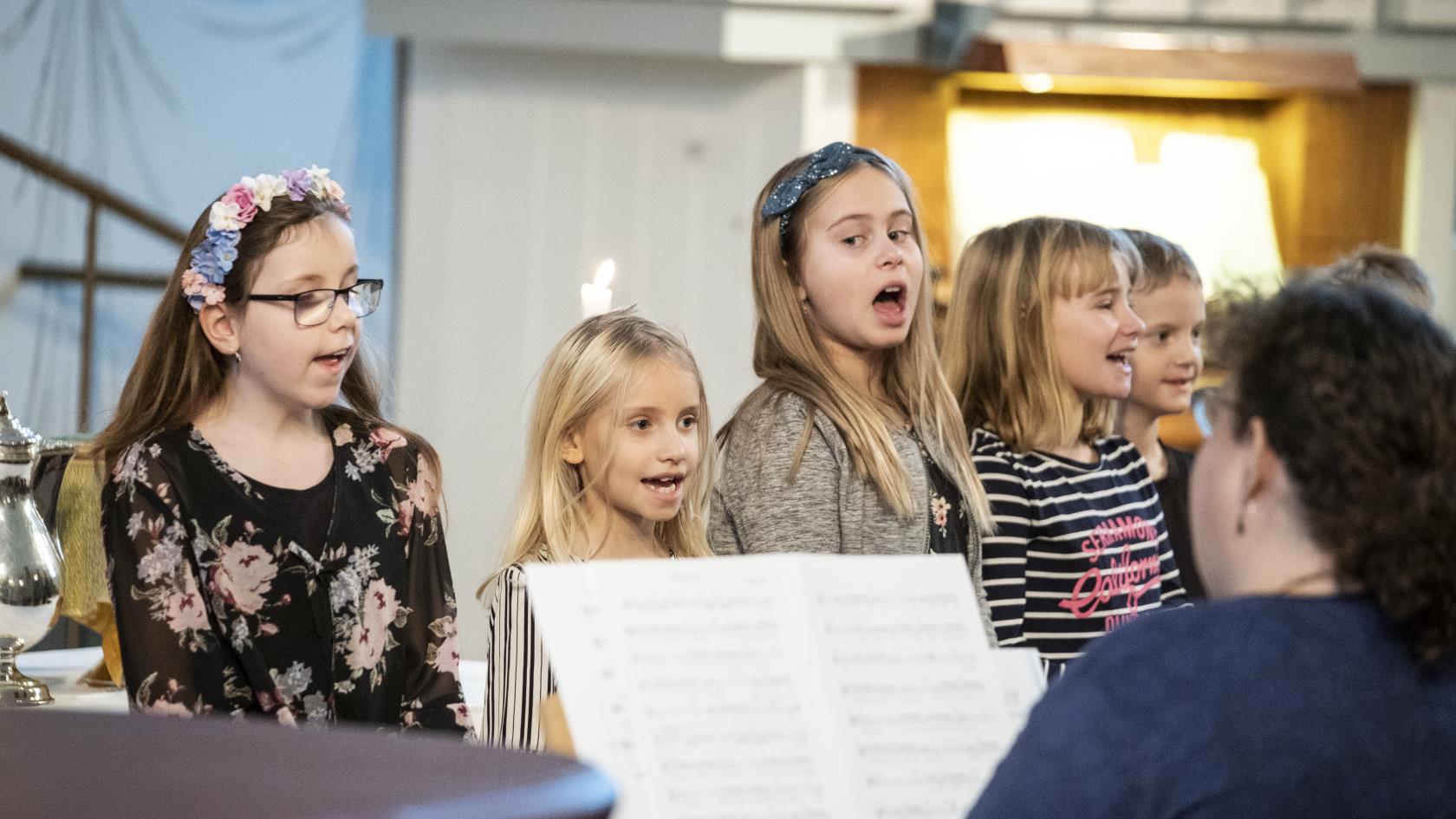 Några barn står bredvid varandra och sjunger framme i kyrkan. En kvinna kompar på piano.