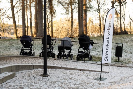 Några barnvagnar står parkerade i en park. En beach-flagga med Svenska kyrkans logga står bredvid.
