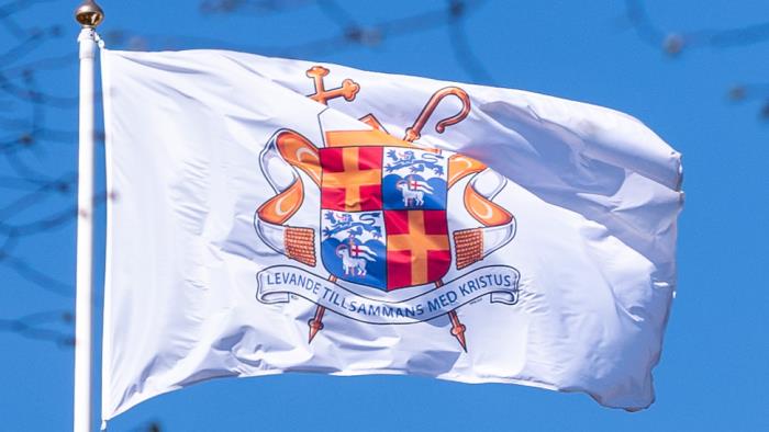 En vit flagga med ärkebiskopens vapensköld mot blå himmel.