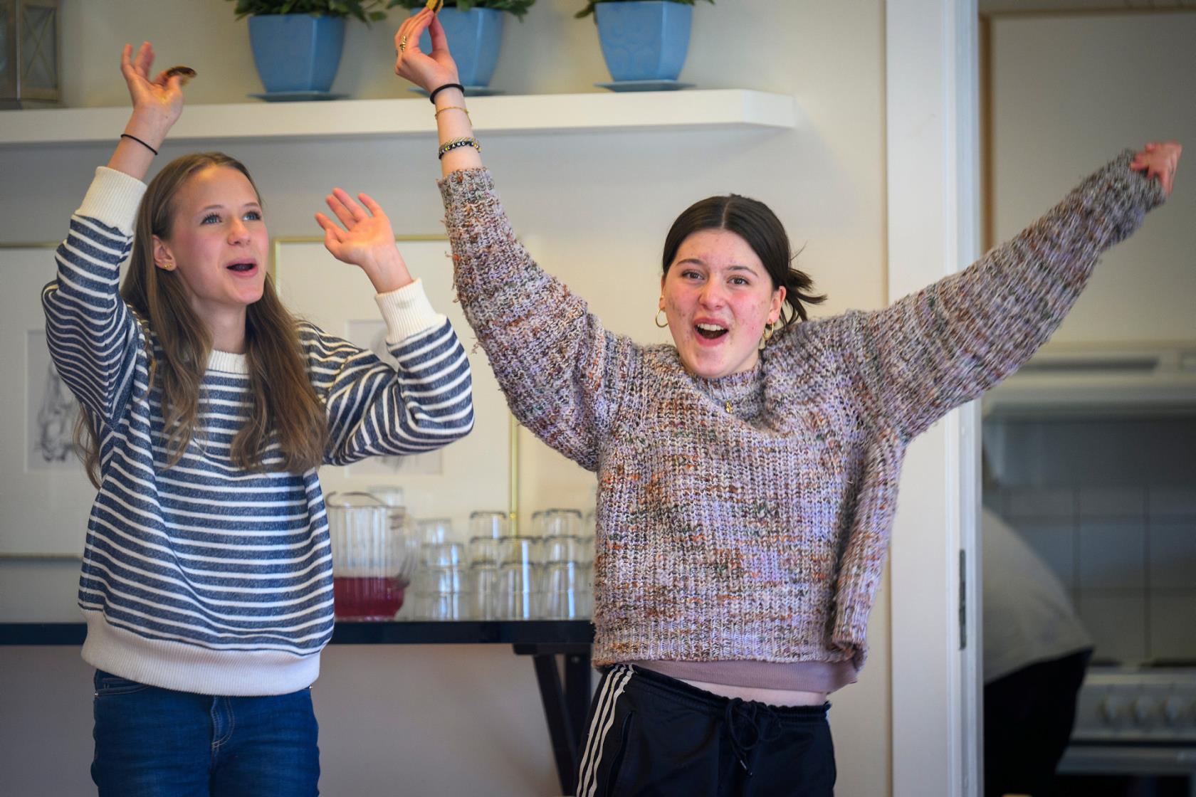 Två unga tjejer dansar glatt och viftar med armarna inomhus.