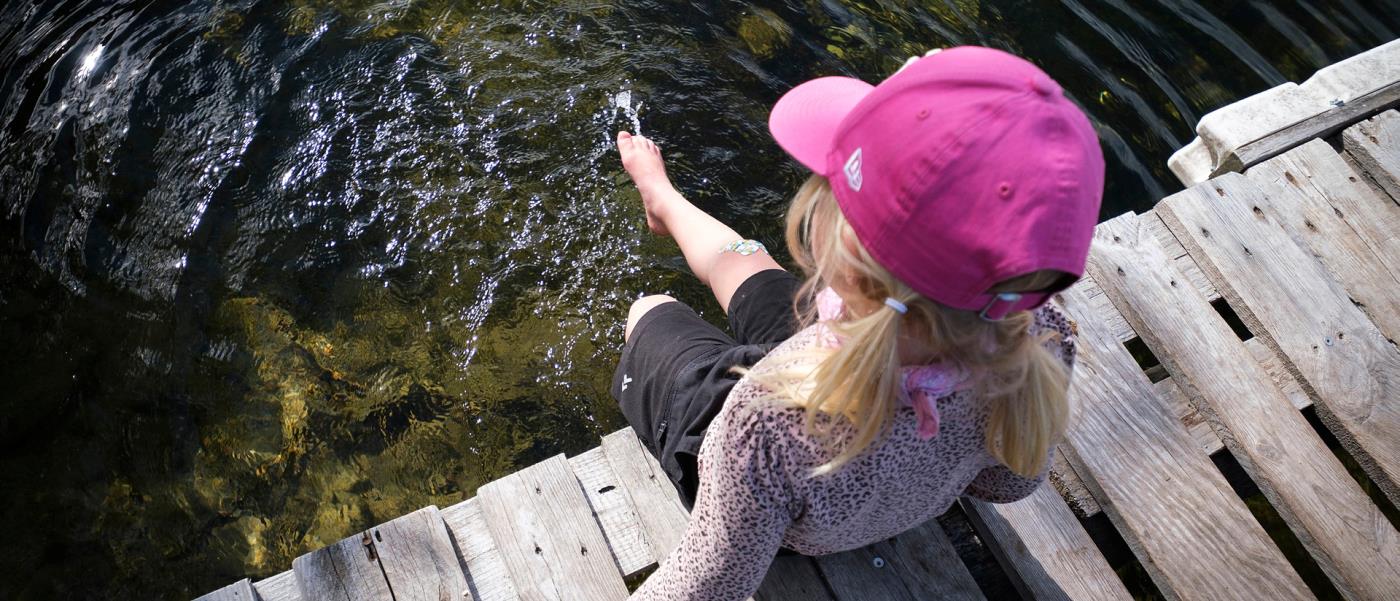 En liten flicka med rosa keps sitter på en brygga och doppar i tårna i vattnet.