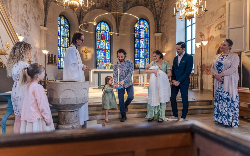 En glad dopfamilj, faddrar och präst står långt fram i kyrkan.