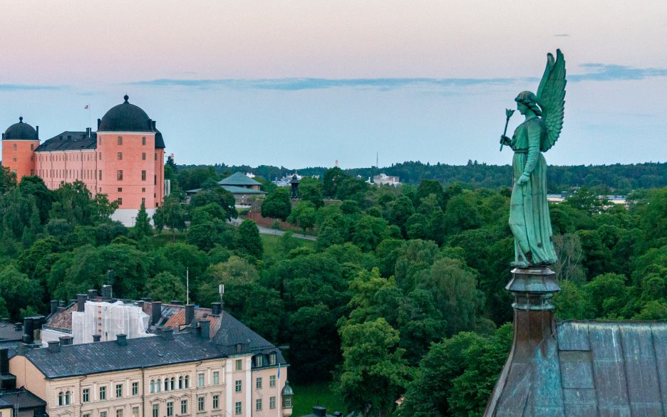 Flygfoto av ängeln längst bak på domkyrkans torn. I bakgrunden ser man grönskande träd.
