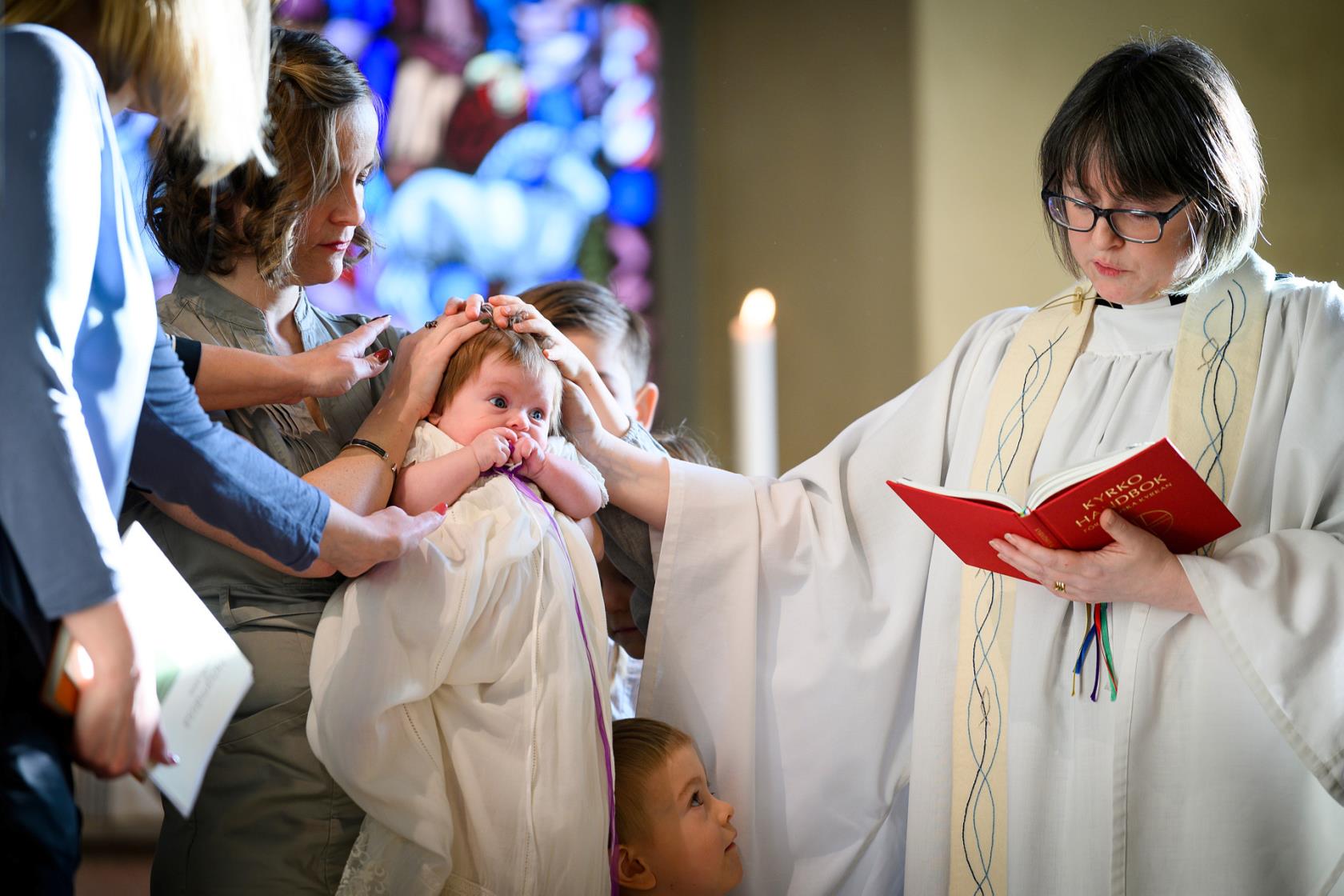 En präst tillsammans med faddrar välsignar ett dopbarn. Liten pojke tittar upp på prästen.