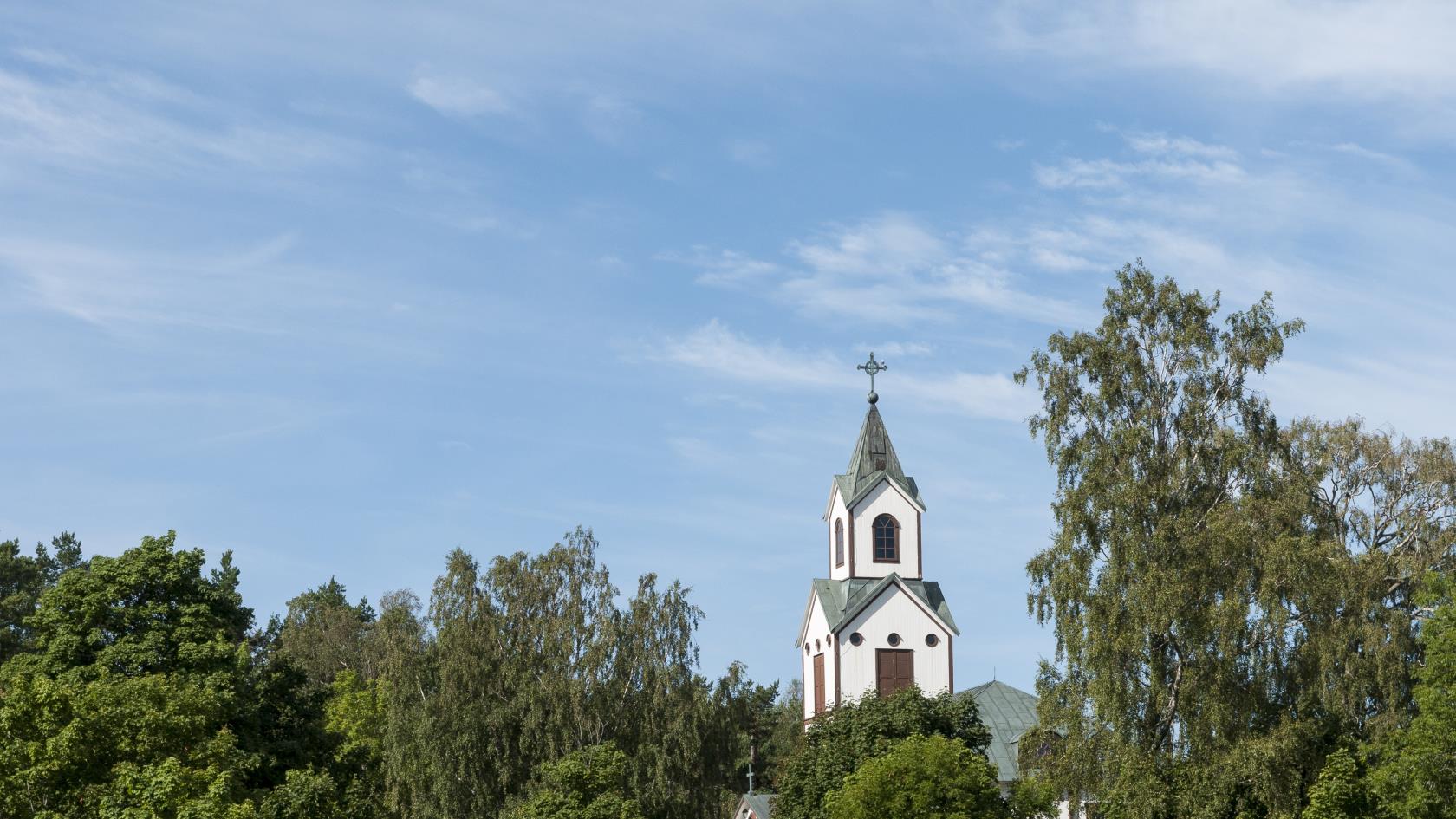 Ett kyrktorn sticker upp bakom trädkronorna mot blå himmel.