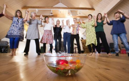 Några kvinnor dansar ringdans runt en skål med vatten och olika frukter som står på golvet.