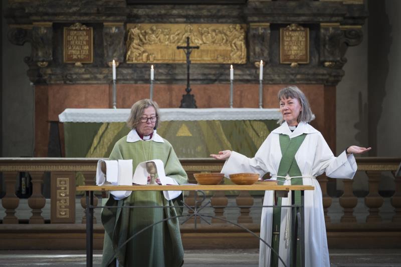 Två kvinnor, den ena präst och den andra biskop, står vid ett uppdukat bord framför ett altare.