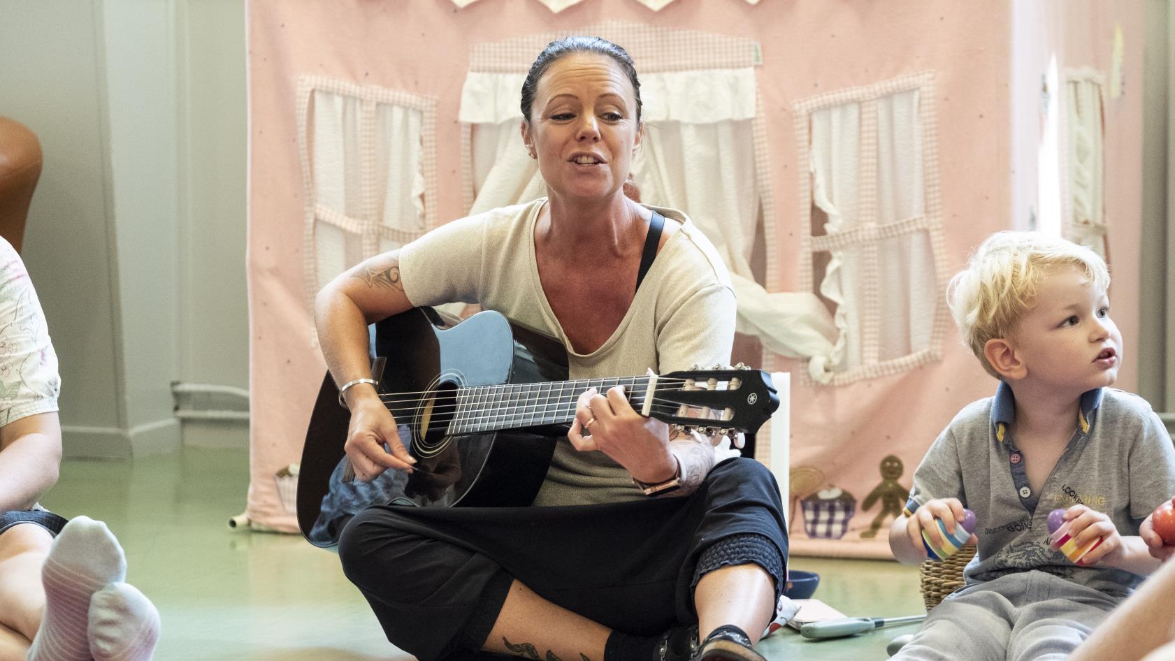 En kvinnlig ledare sitter i en ring på golvet med barn och deras föräldrar. Hon spelar gitarr och sjunger.