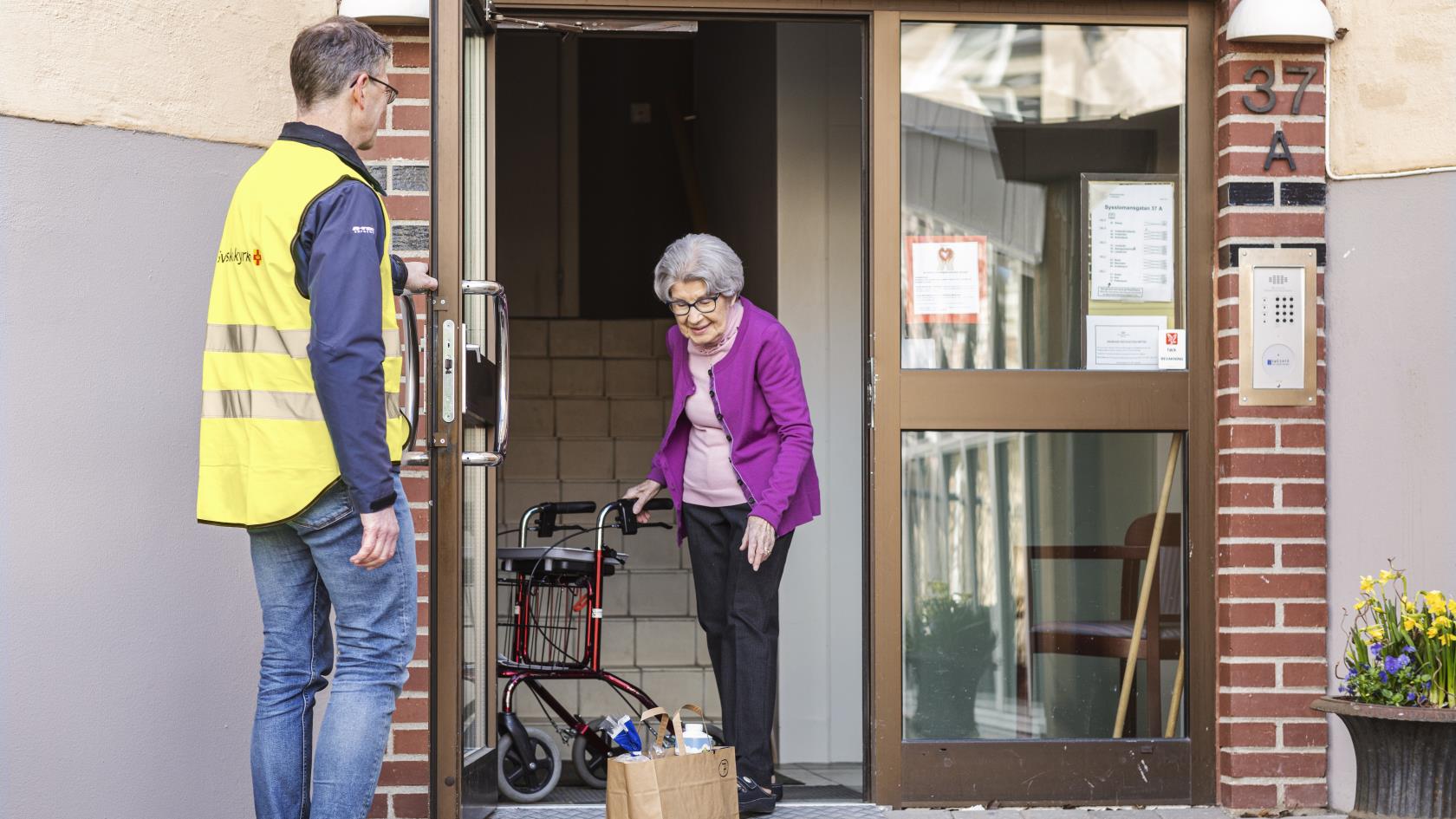 Man från Svenska kyrkan möter en äldre dam i porten till hennes hus och lämnar en matkasse.