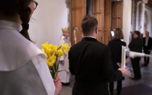 En kvinna i vit särk står med en bukett påskliljor i absiden till Uppsala domkyrka, redo att gå in tillsammans med några andra.