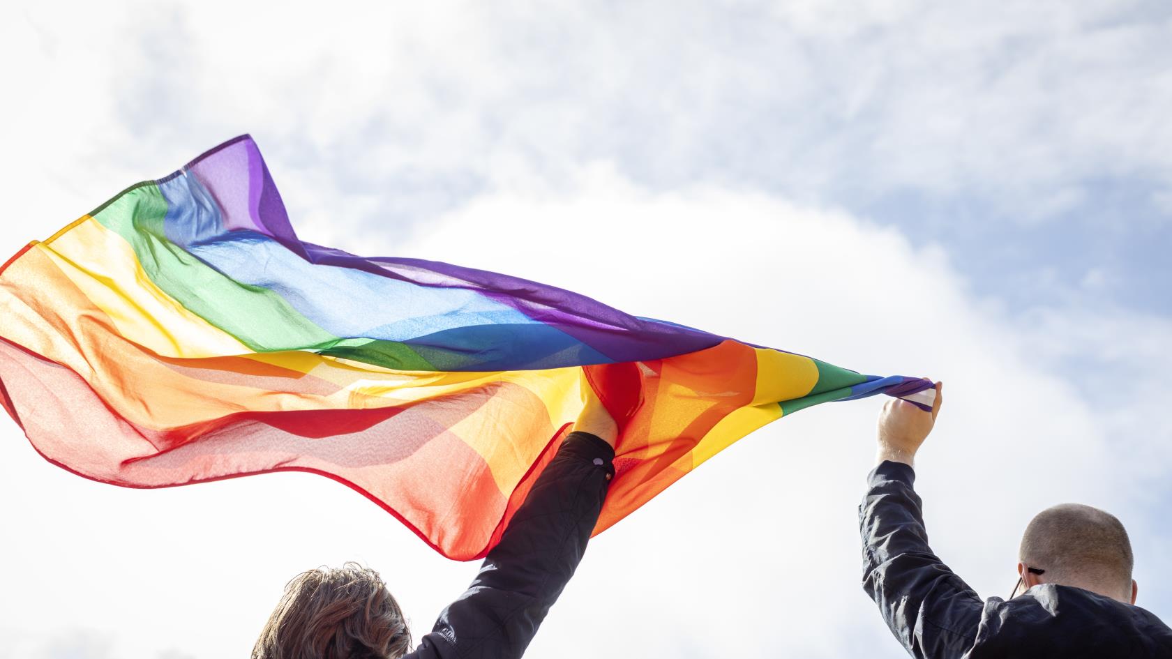 Två personer håller upp regnbågsflaggan mot himlen.