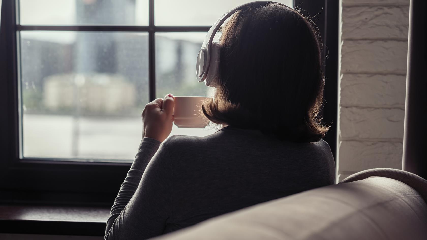 En kvinna med hörlurar sitter i en soffa med en kopp kaffe och tittar ut genom fönstret.