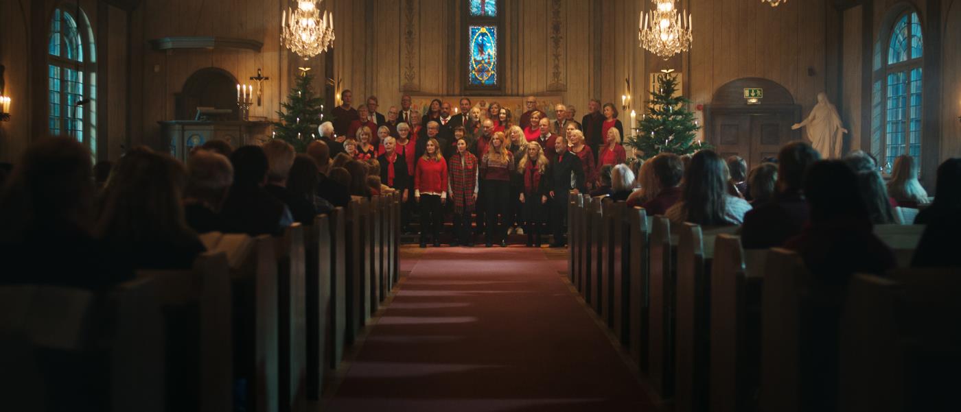Mittgången i en fullsatt kyrka. Längst fram står en kyrkokör i julkläder mellan två julgranar och sjunger.