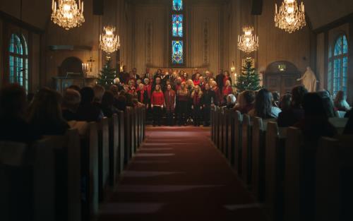 Mittgången i en fullsatt kyrka. Längst fram står en kyrkokör i julkläder mellan två julgranar och sjunger.