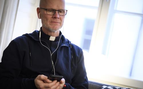 En manlig präst sitter i en soffa med en mobiltelefon i handen och hörlurar i öronen.