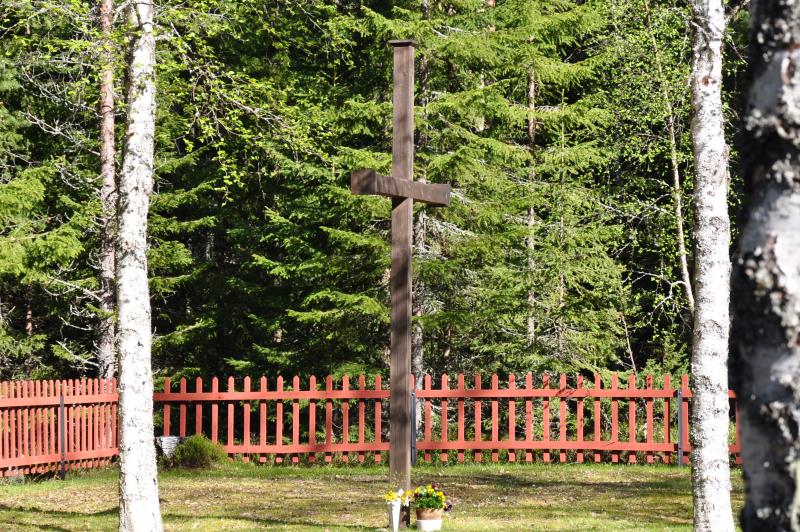 Det mörka korset på Björkbergs kyrkogård omges av vitstammiga björkar och ett rött trästaket. 