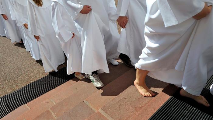 Konfirmander som går uppför kyrktrappan på väg in i kyrkan och sin konfirmation. Fokus ligger på fötterna som är med och utan skor. De håller hand och är iklädda vita kåpor.