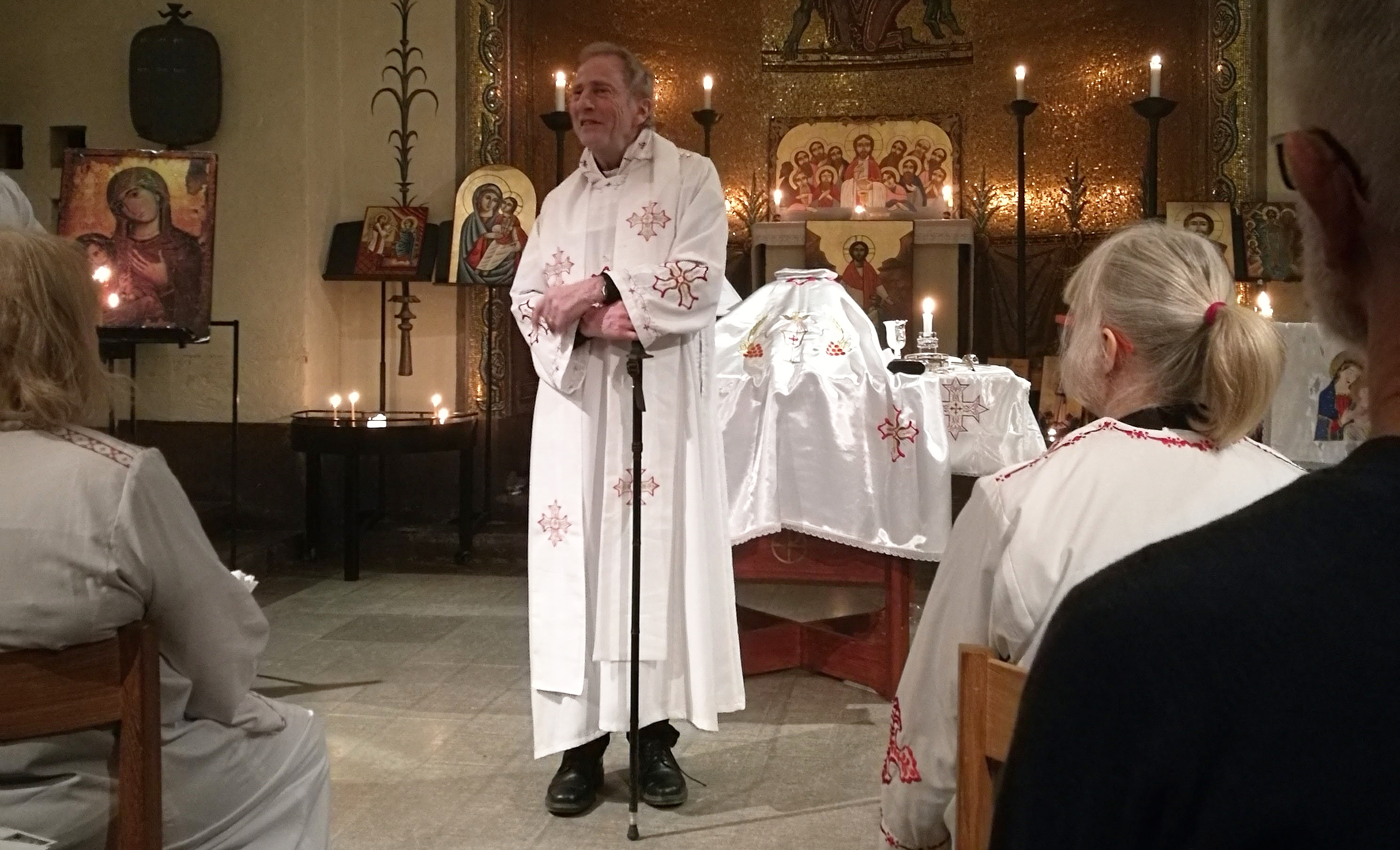 En person i prästdräkt står och framför hen sitter ett antal personer och bakom hen är ett altare. Det är tända ljus på flera  håll i kyrkrummet. 