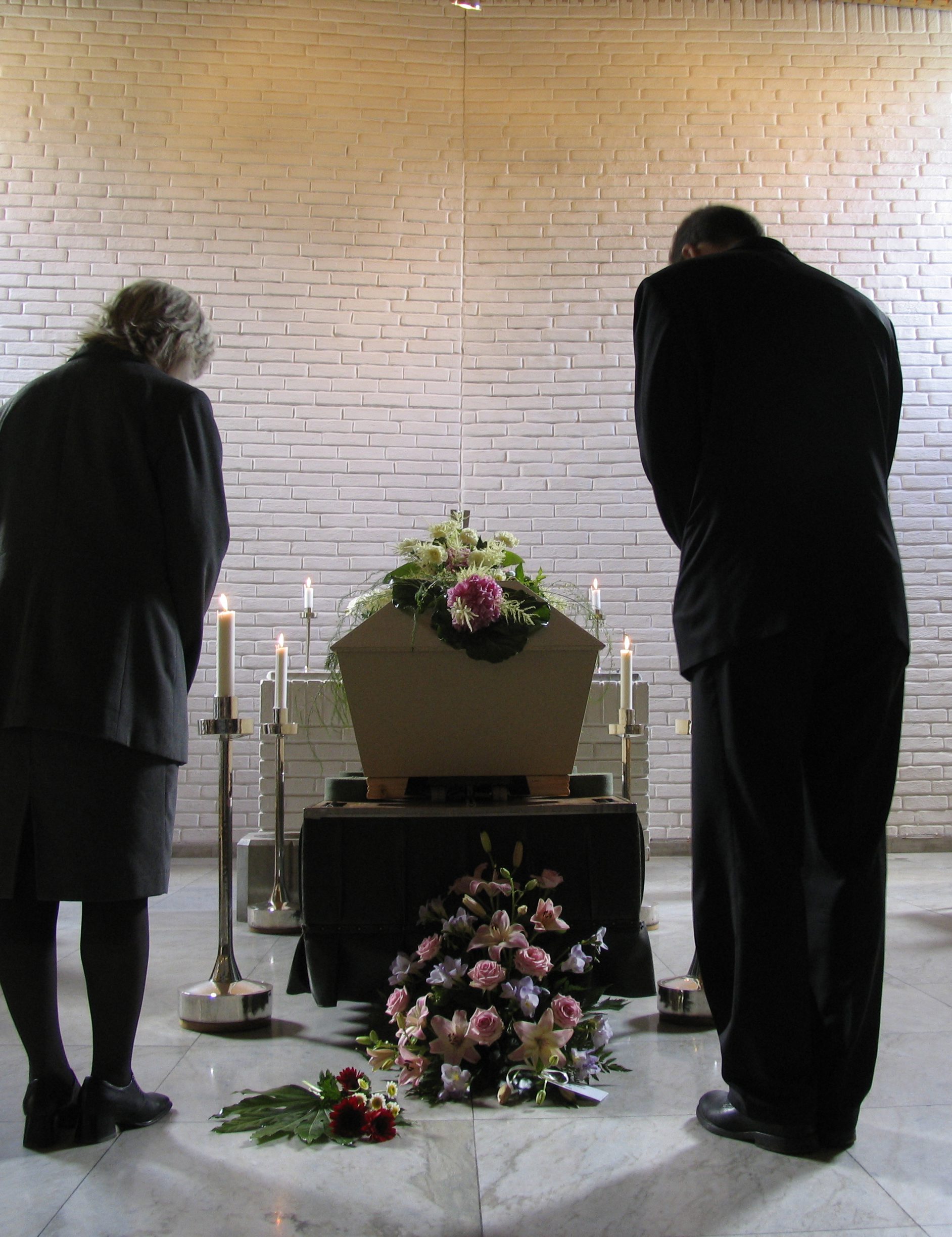 En svartklädd kvinna och en svartklädd man bugar framför en kista med blommor på.