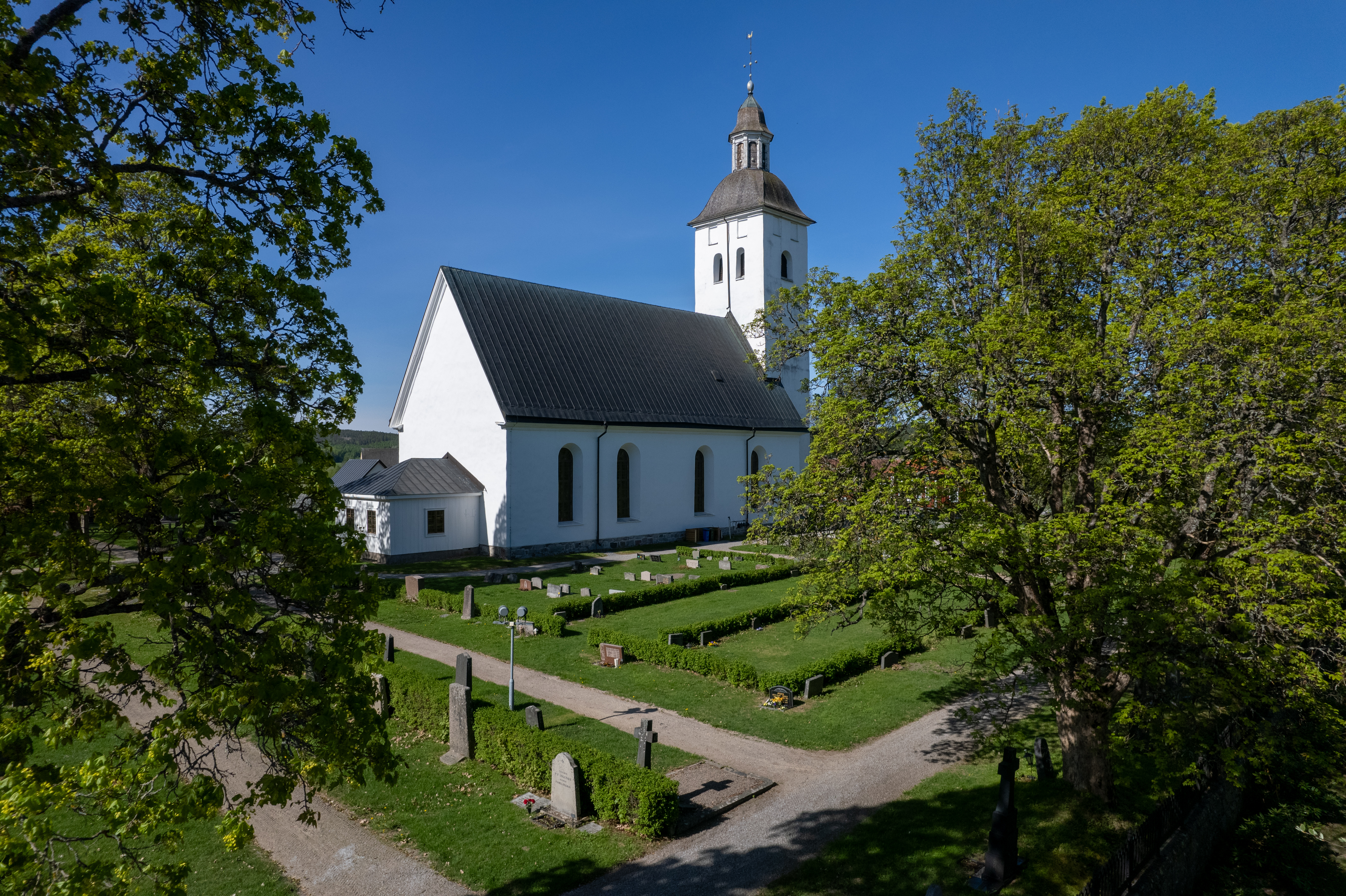 En drönarbild på Hälsingtuna kyrka omgiven av gröna lövträd och kyrkogård