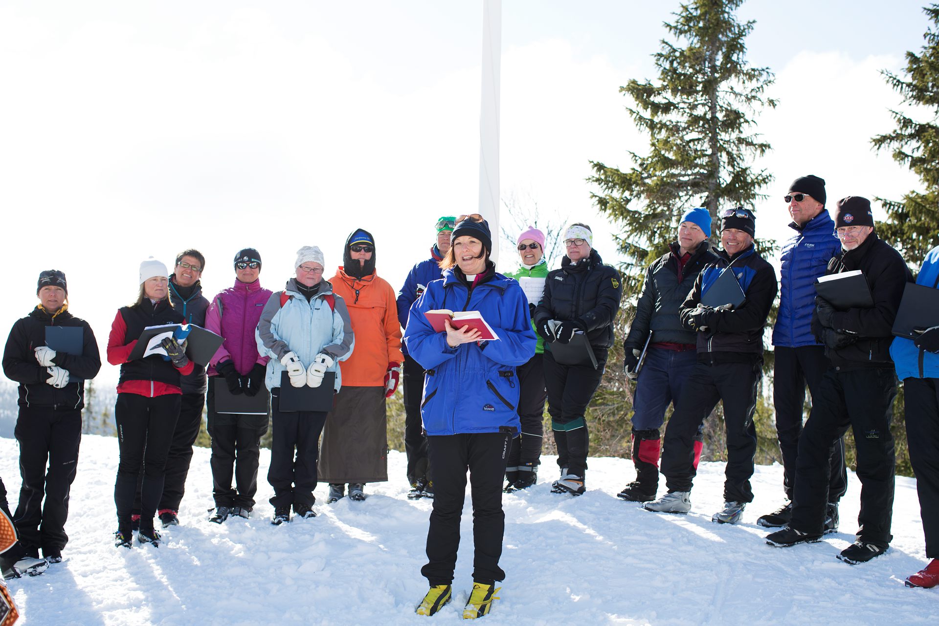 Utomhus en solig vinterdag. Prästen Karin Härjegård klädd i vinterkläder och skidpjäxor. Hon håller en psalmbok i sin hand och flankeras av ett tiotal personer också de klädda för vinter och skidåkning.