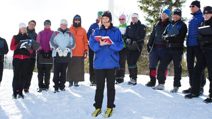 Utomhus en solig vinterdag. Prästen Karin Härjegård klädd i vinterkläder och skidpjäxor. Hon håller en psalmbok i sin hand och flankeras av ett tiotal personer också de klädda för vinter och skidåkning.