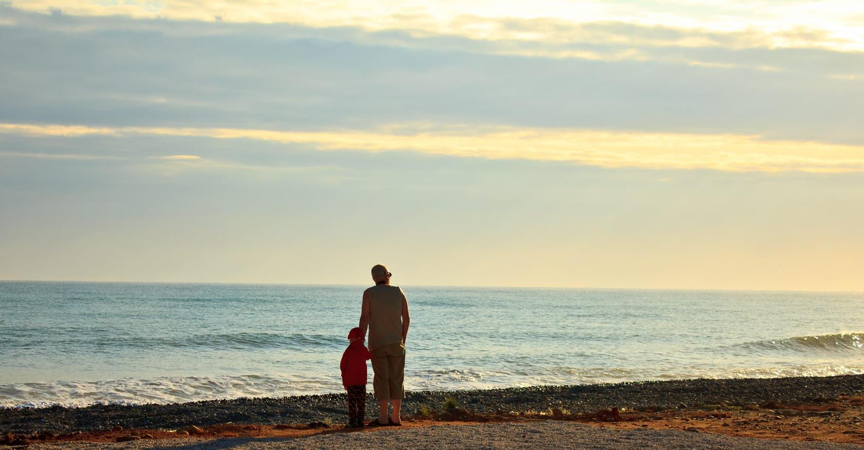 En man och ett barn står på stranden och tittar ut över havet. Vågor rullar in.
