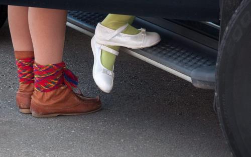 Två par fötter skymtar bakom en bildörr. Det ena paret är klädda i näbbskor med frägranna vävda band.