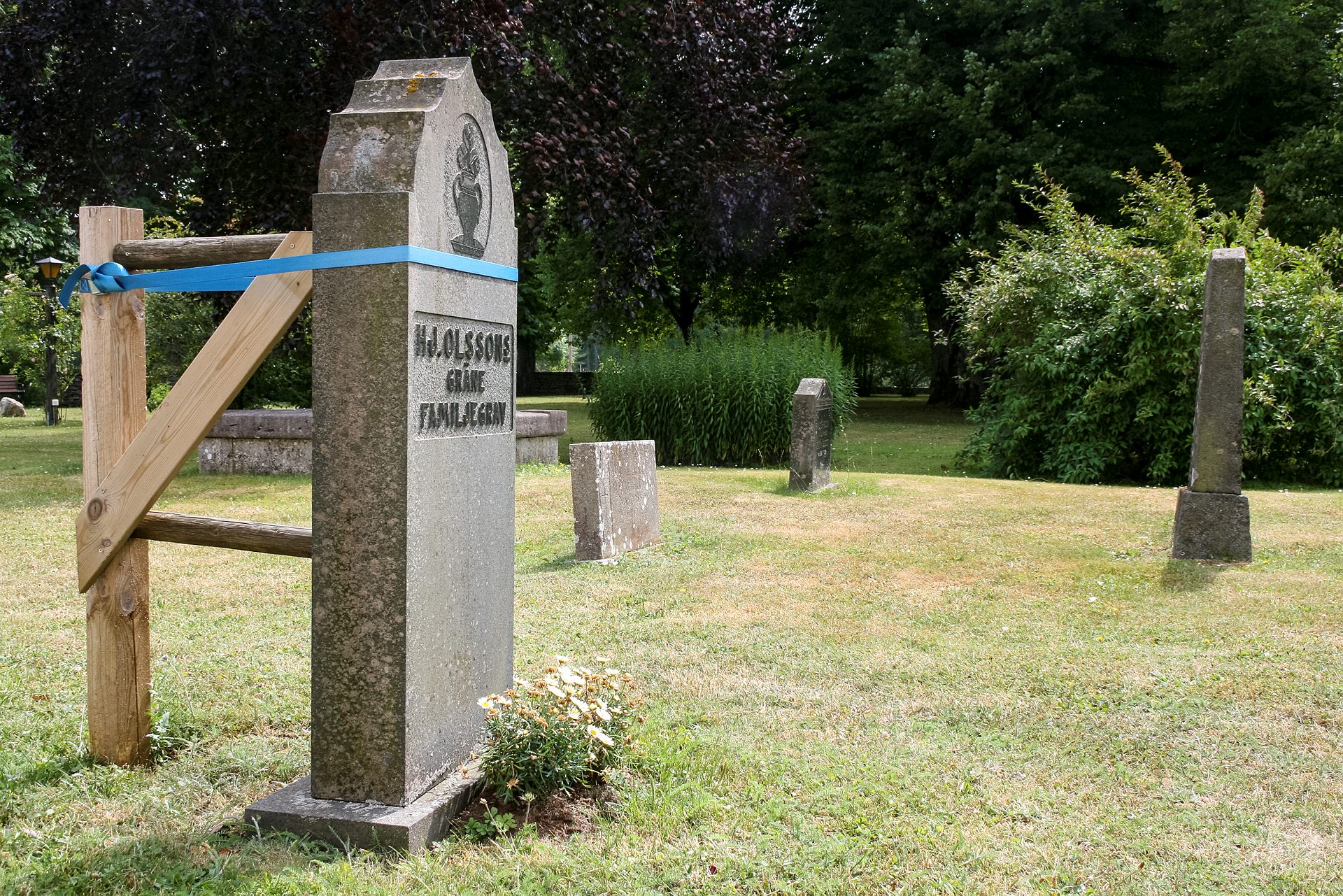 En gravsten stöttas med hjälp av en påle och spännband. Några andra gravstenar syns i bakgrunden.