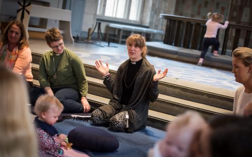 På en blå filt på kyrkgolvet nedanför koret sitter en kvinnlig präst. Vid hennes vänstra sida sitter en kvinnlig diakon. I bakgrunden kan man se en liten flicka på väg att klättra på knäfallet framför altaret. på bilden syns även fler små barn och deras mammor.