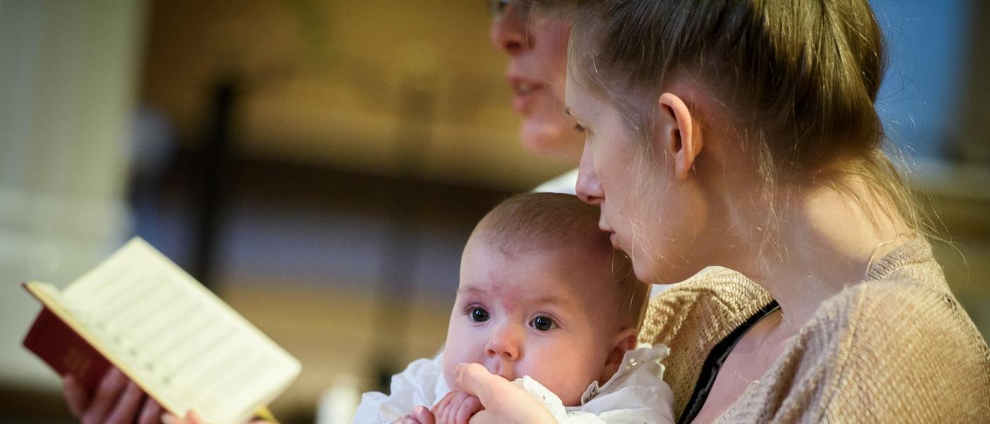 Närbild på föräldrar med bebis i dopdräkt. De håller en psalmbok framför sig.