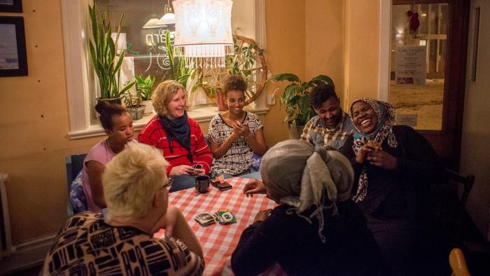 Ett brokigt sällskap av olika nationalitet och ursprung spelar sällskapsspel och skrattar kring ett bord med rutig duk.