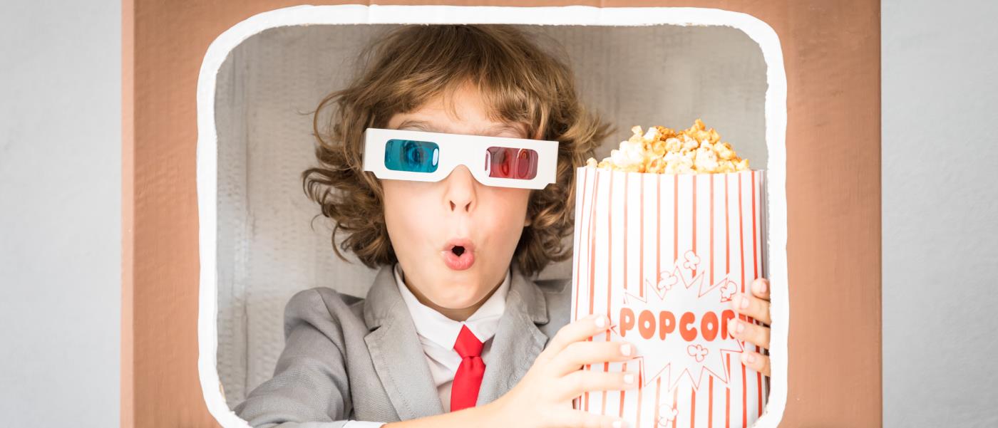 Pojke som tittar ut genom en tv-ruta. Han har 3d-glasögon på sig och håller i en rödranding förpackning med popcorn.