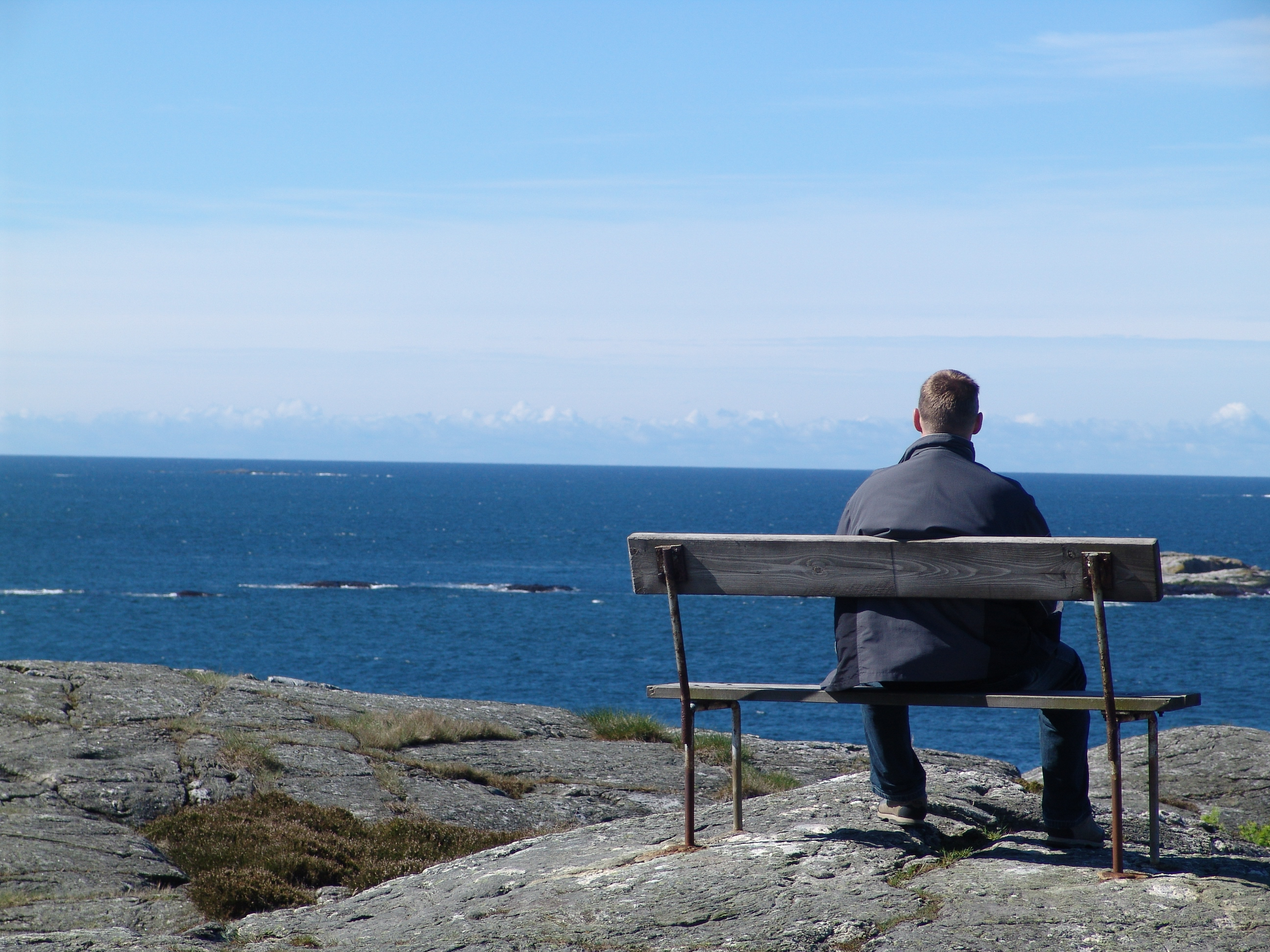En ensam man sitter på en bänk på en klippa och ser ut över havet