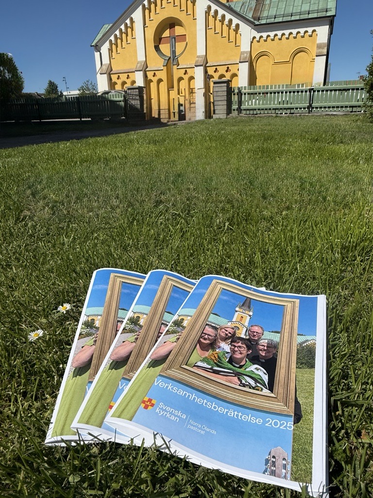 Tre häften med bild på kyrka och personal ligger i gräset framför kyrkan