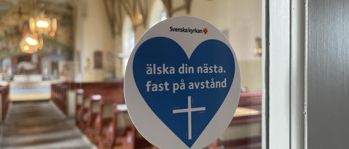 Ett klistermärkeshjärta med texten "Älska din nästa fast på avstånd" sitter på en fönsterruta i en kyrka