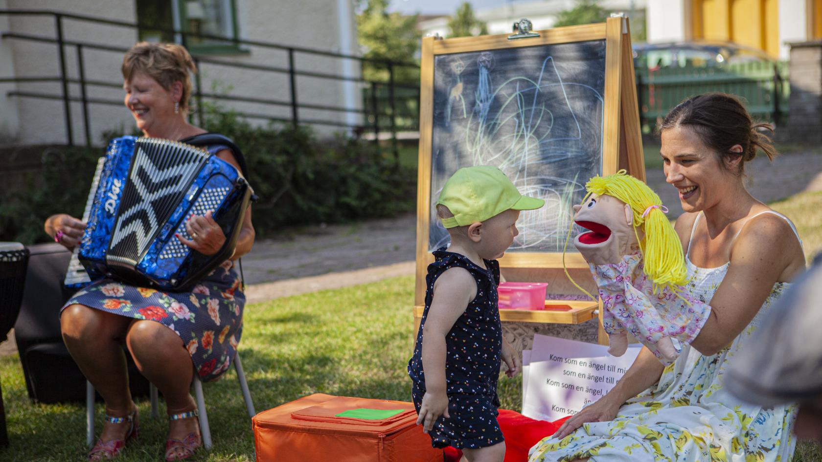 Handdockan Bibbi, barnledaren Julia och kantor Helen som spelar dragspel pratar med ett litet barn utomhus en sommardag