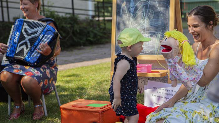 Handdockan Bibbi, barnledaren Julia och kantor Helen som spelar dragspel pratar med ett litet barn utomhus en sommardag