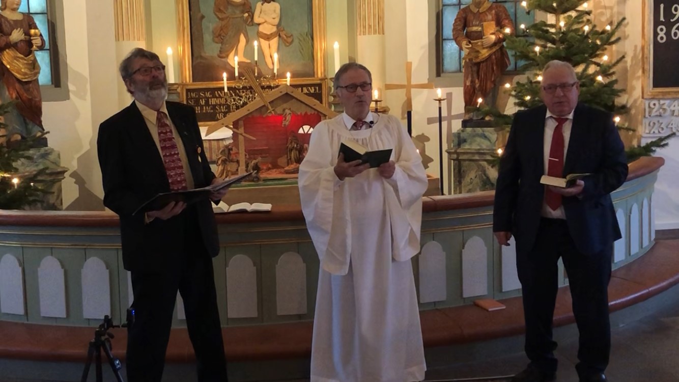 Tre sjungande herrar framför en julsmyckad kyrka